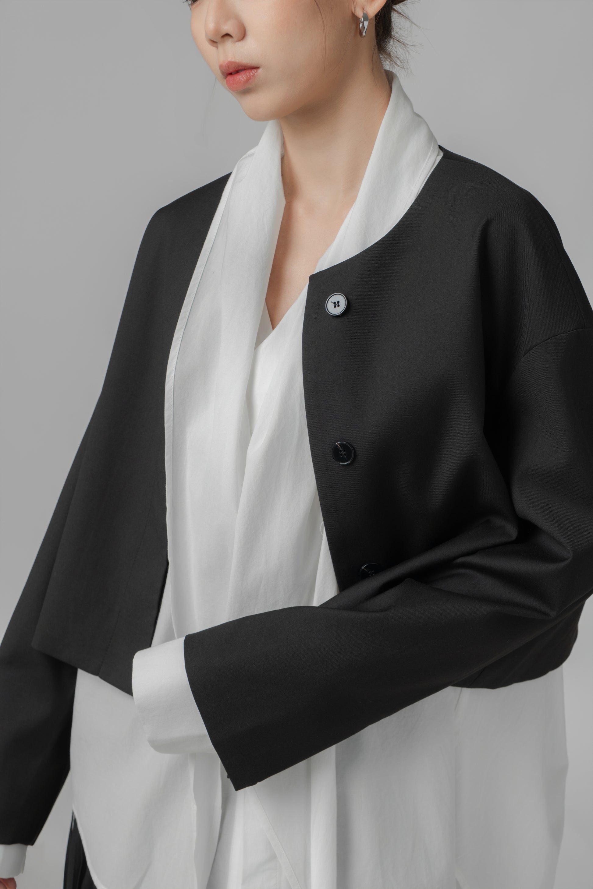 Woman wearing a black cropped, boxy collarless jacket with front buttons over a white shirt 