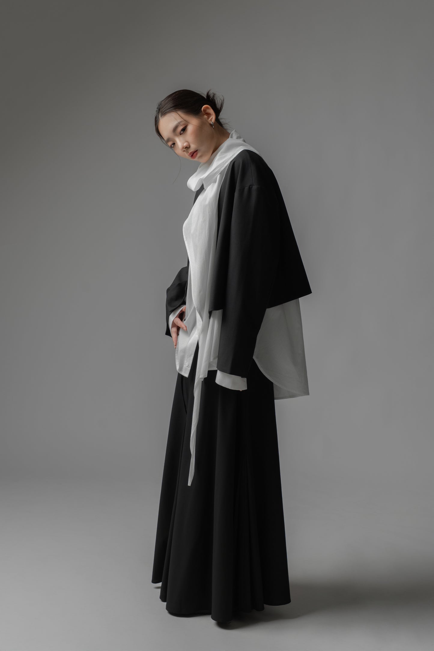 Woman wearing a black cropped, boxy collarless jacket over a white shirt with scarf collar and black skirt 