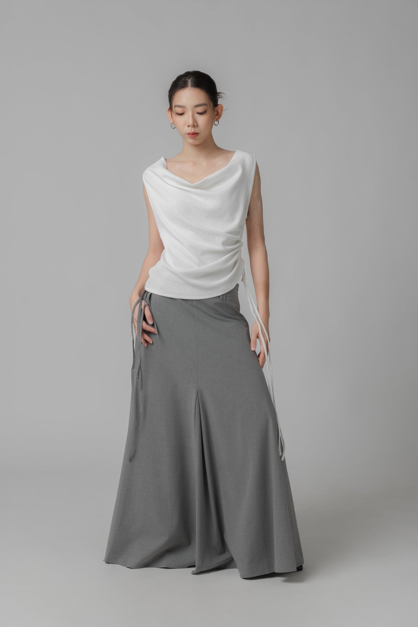 Woman wearing a white sleeveless wide-neck top with adjustable side string, paired with a grey skirt