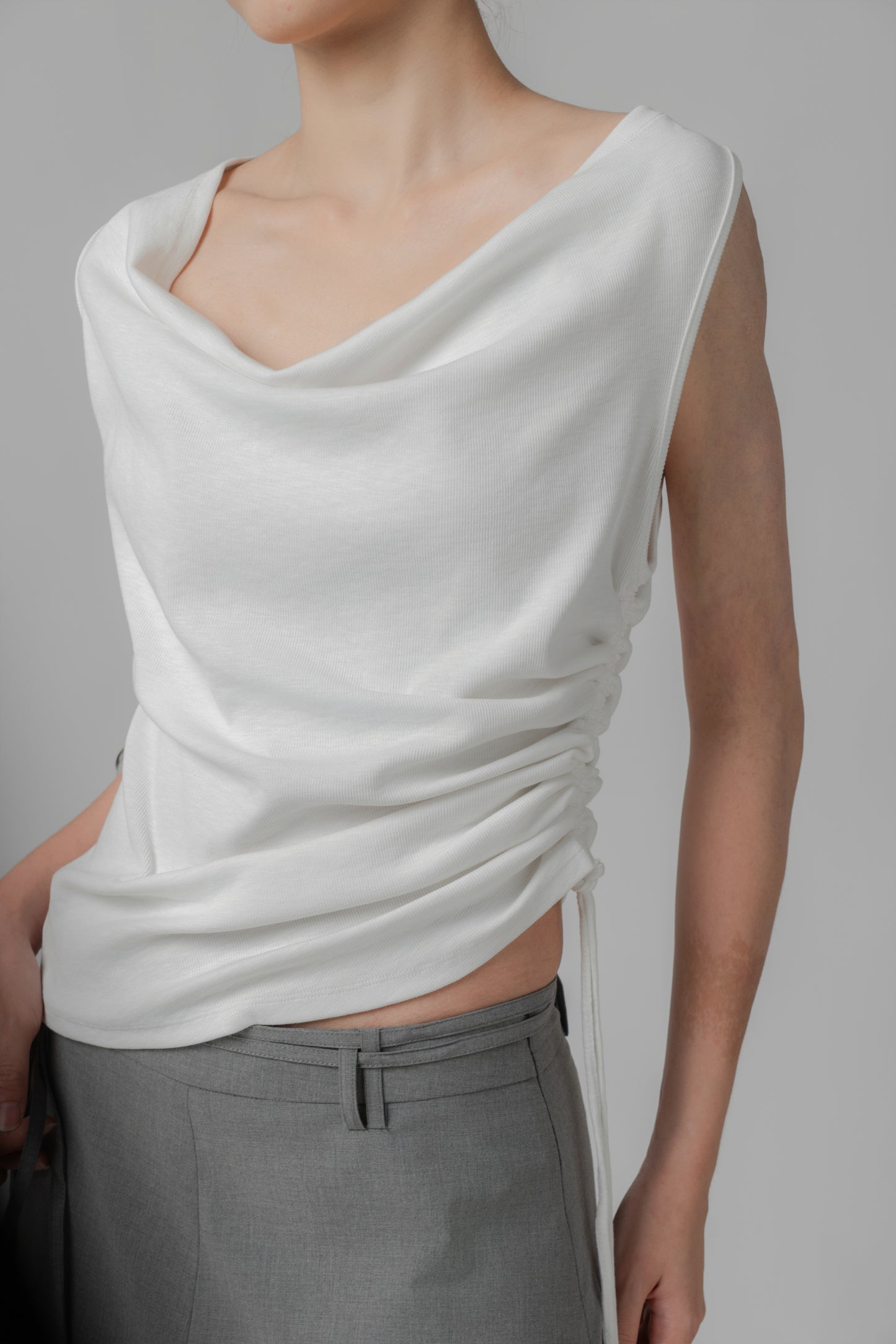 Woman wearing a white sleeveless wide-neck draped top with adjustable side string, paired with a grey skirt