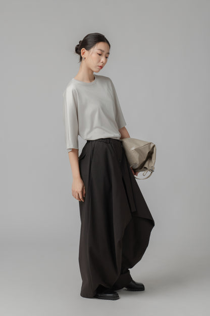 Person wearing a light gray asymmetric T shirt and black wide-leg pants with a leather bag on a plain background
