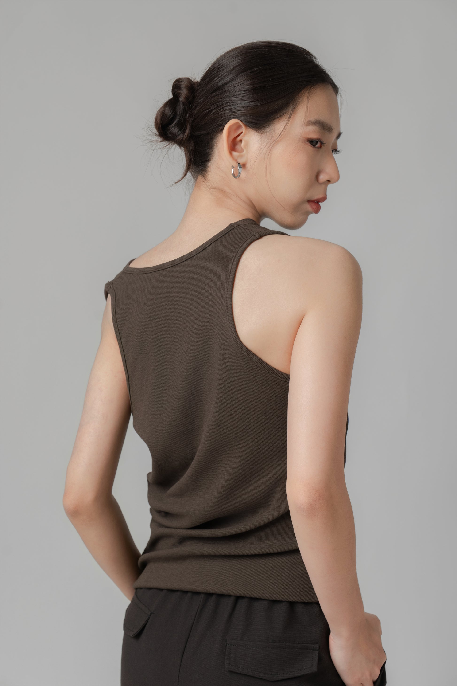Woman showing the back of a dark brown sleeveless one-shoulder draped top with adjustable side string, paired with black wide leg trousers