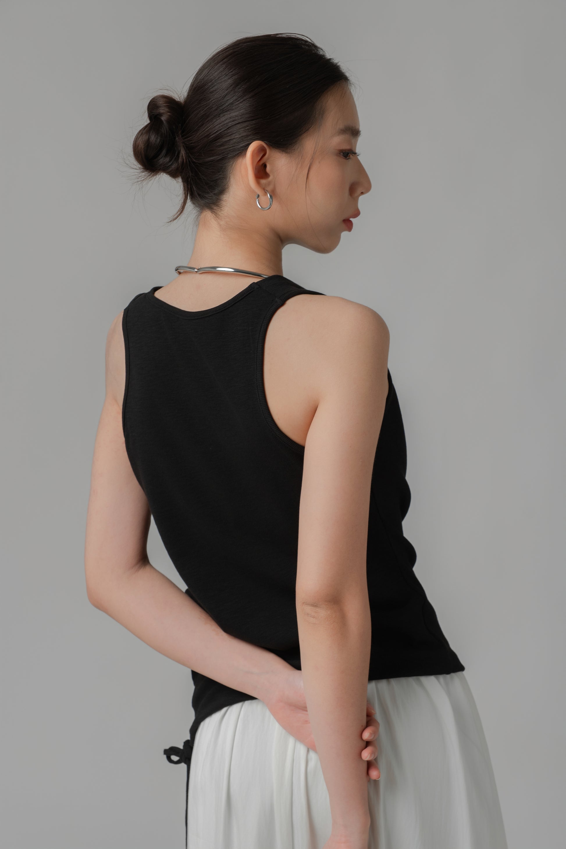 Woman showing the back of a black V-neck sleeveless top with adjustable side string, paired with a white skirt