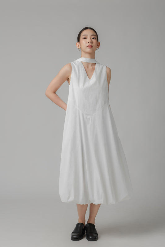 Woman wearing a white sleeveless bubble hem dress with scarf detail against a gray background