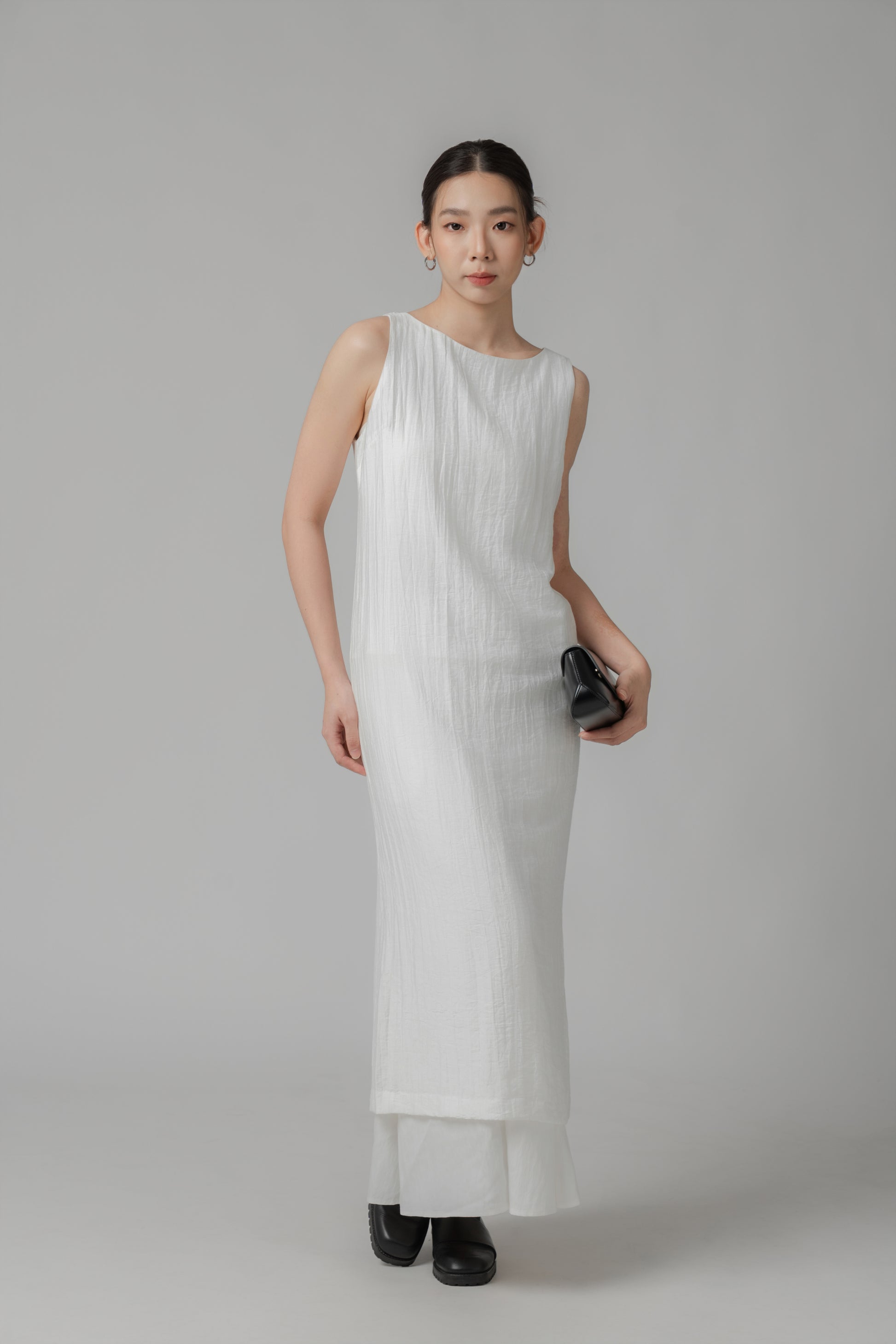 Woman wearing a white sleeveless crinkled column dress layered over a skirt and styled with a black leather bag